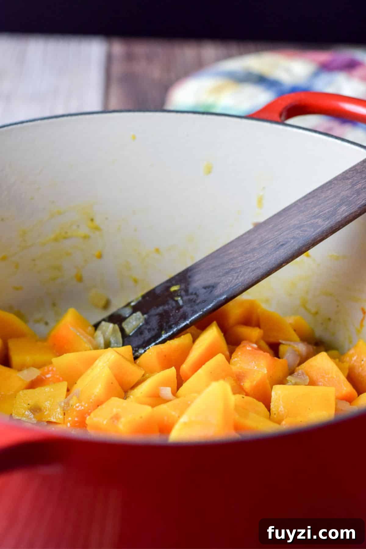 Creamy Roasted Butternut Squash Soup Recipe 5 Cut squash cooked in chicken broth with some onions in a red pot and a wooden spoon in the pot