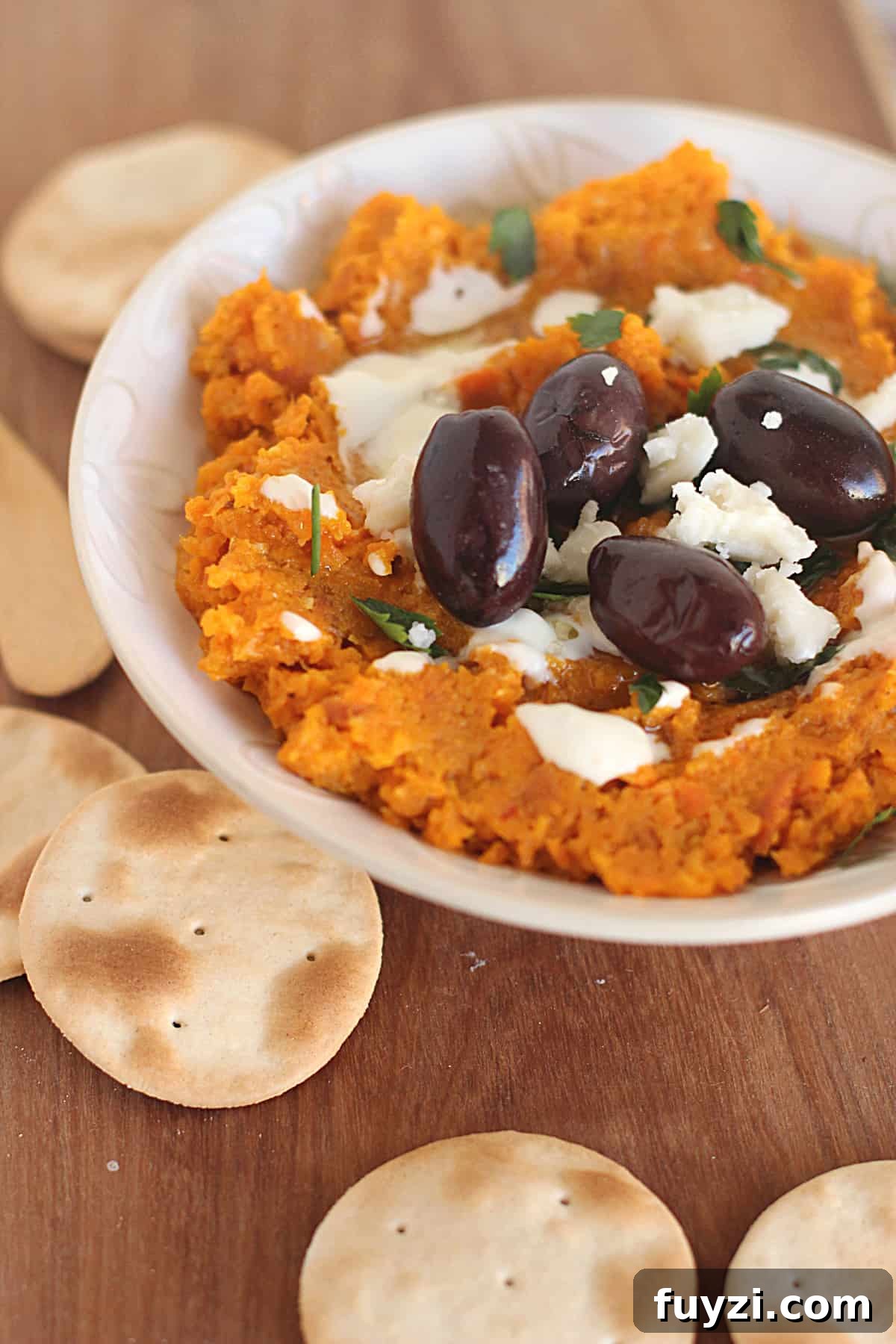 On a wooden board a white bowl with carrot dip and black olives surrounded by crackers