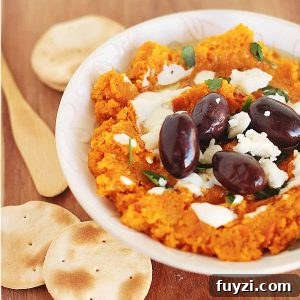 Creamy carrot dip with black olives and feta cheese in a white bowl with crackers.