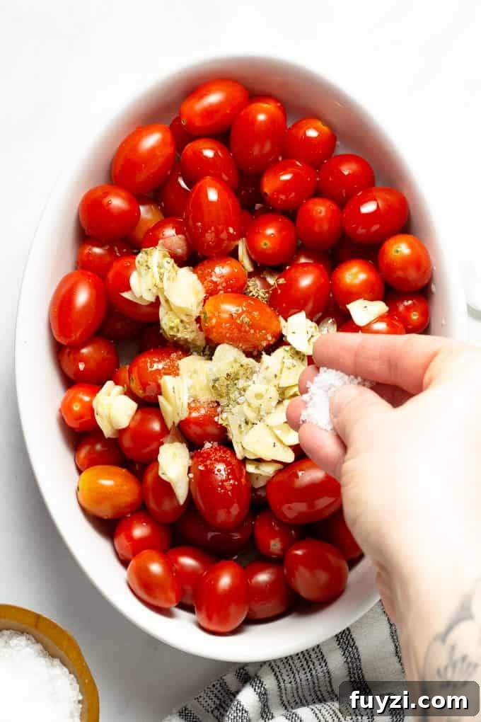 Overhead shot of a white baking dish filled with tomatoes, garlic, oregano and olive oil with a hand sprinkling Kosher salt over it