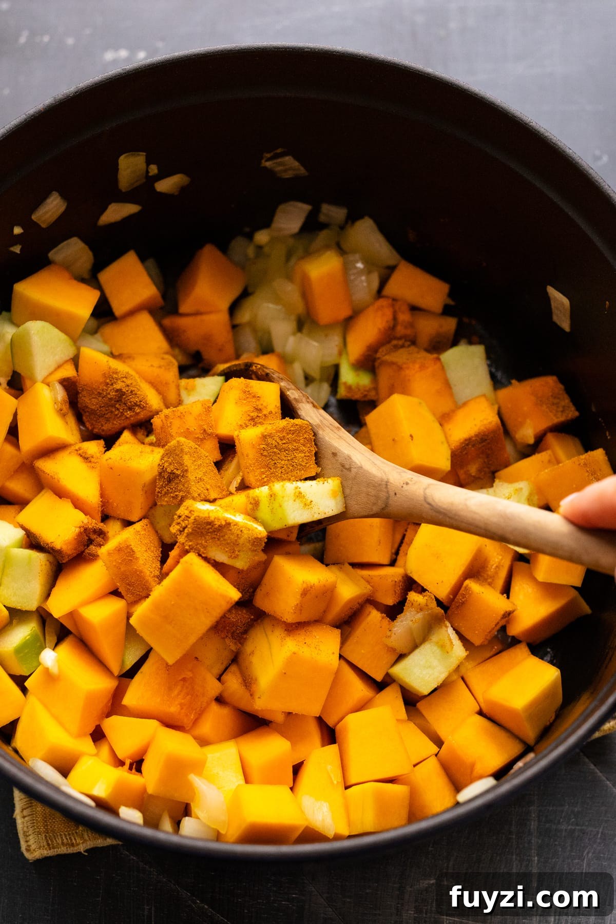 Curried Butternut Squash Soup Recipe 5 A wooden spoon stirring diced butternut squash and apples in a black cast iron pot.