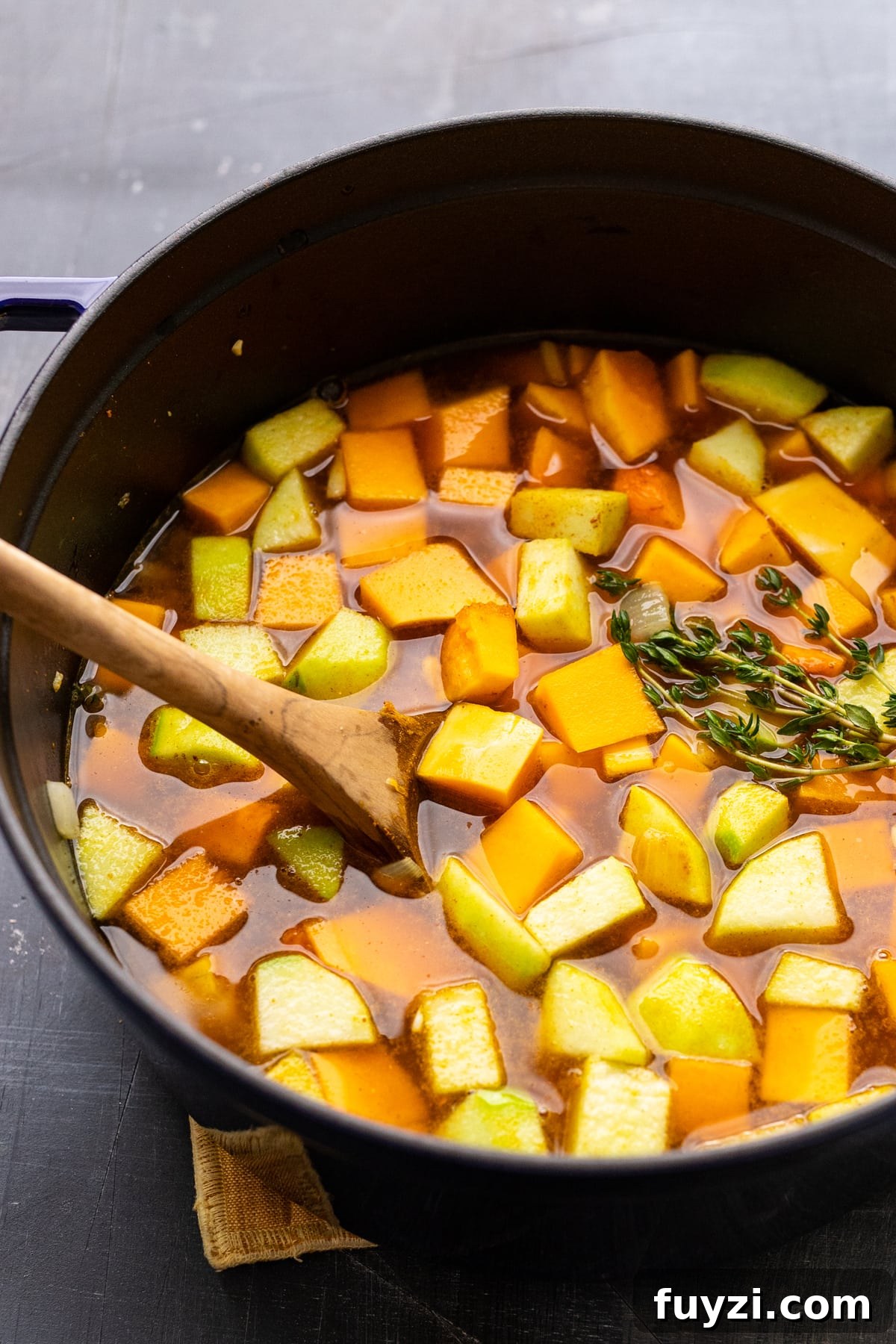 Curried Butternut Squash Soup Recipe 7 A wooden spoon stirring cubed apples and butternut squash in broth.