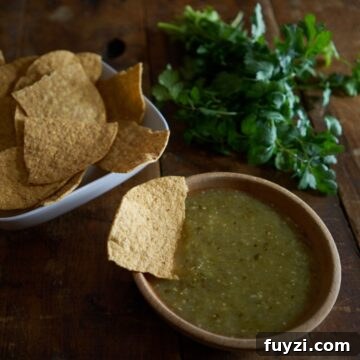 A wooden bowl of tomatillo salsa verde with a chip in it. Fresh cilantro and a bowl of chips are in the background.