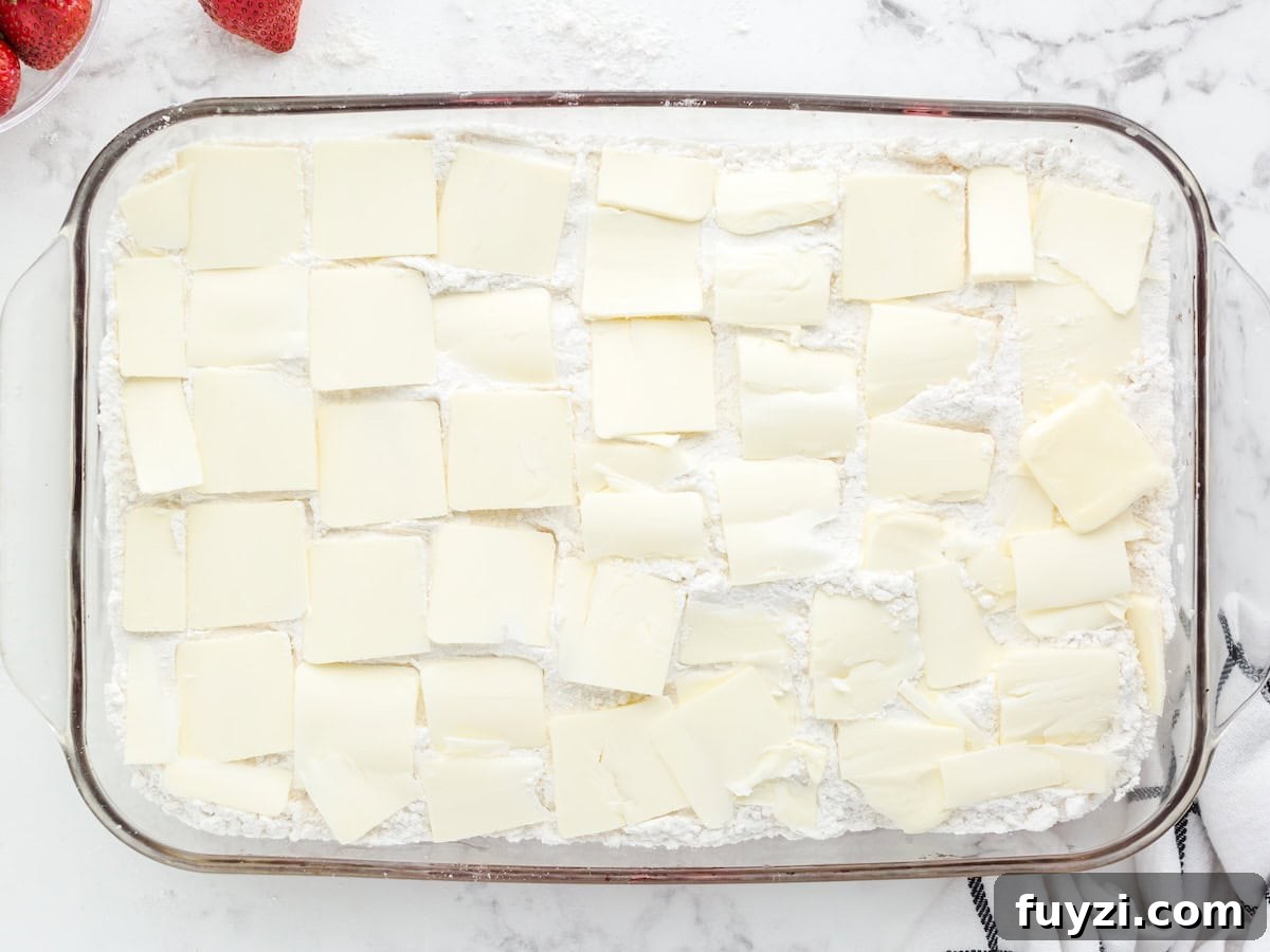 A glass baking dish filled with a white powdery layer, topped evenly with overlapping slices of butter, hints at the start of a delicious Strawberry Dump Cake Recipe; strawberries and a striped towel are nearby.