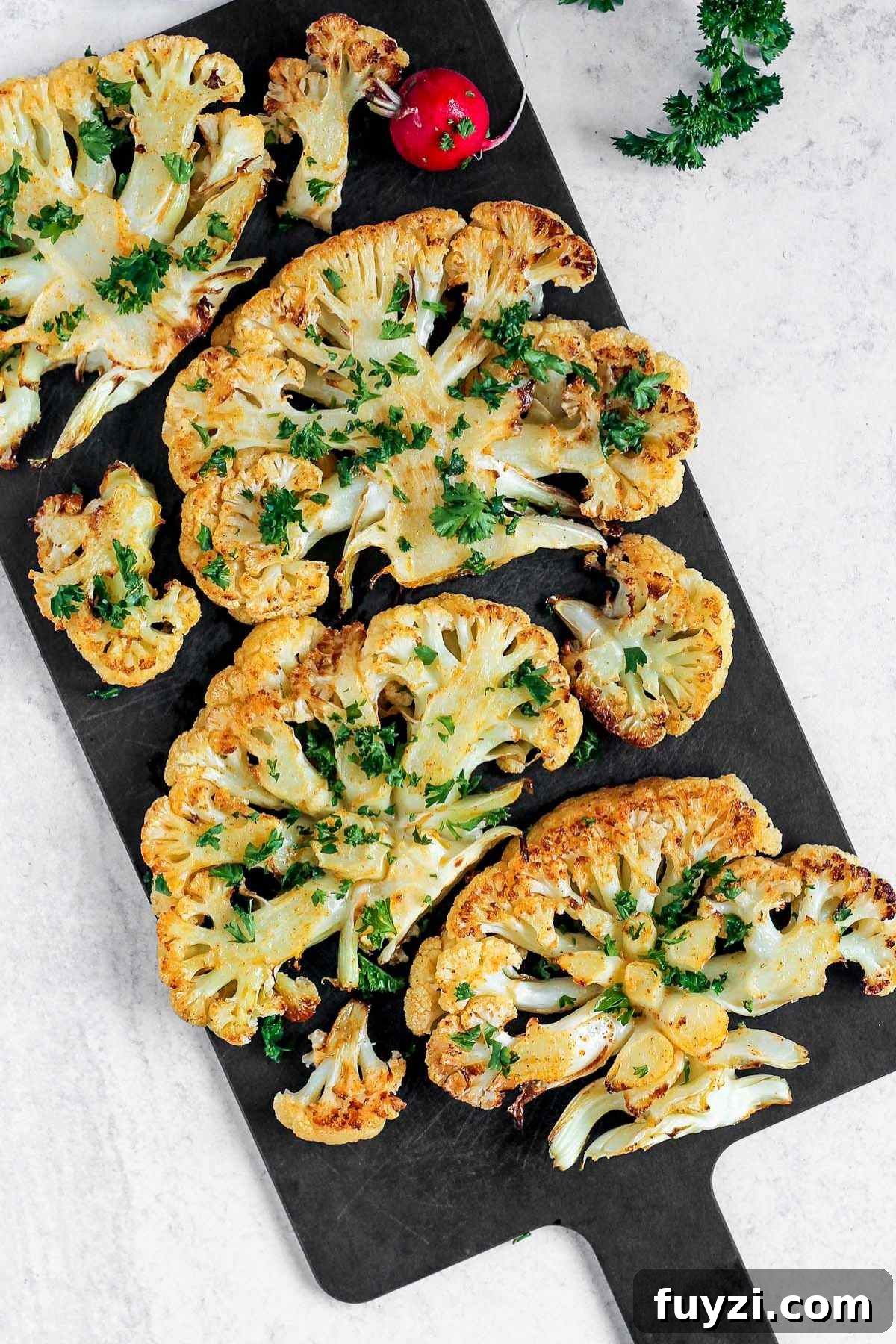 Vegan Roasted Cauliflower Steaks with Garlic-Herb Sauce 2 Roasted cauliflower steaks sprinkled with fresh parsley on a black cutting board.