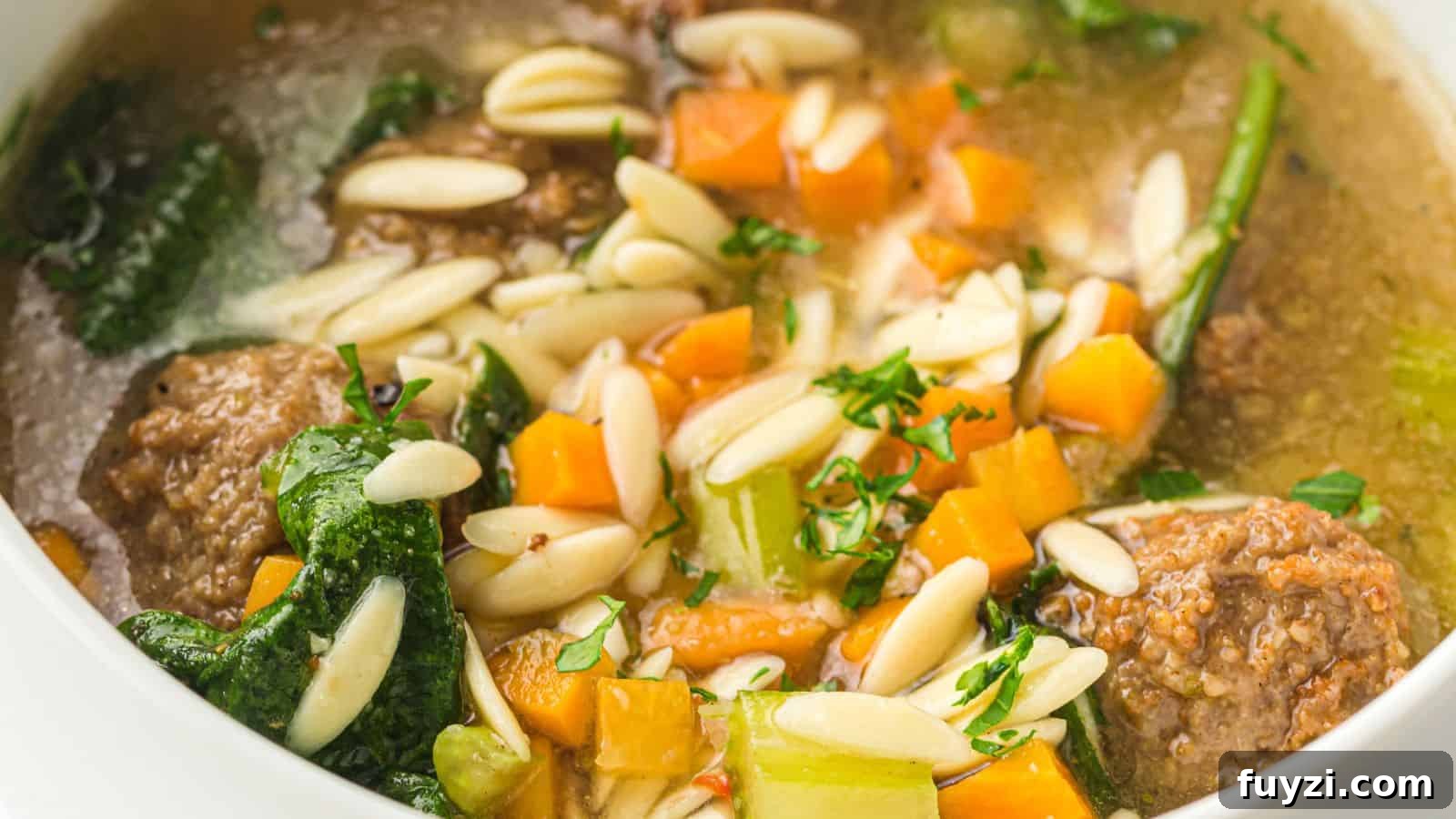 A close-up of soup with meatballs, carrots, celery, spinach, slivered almonds, and fresh herbs in a clear broth.