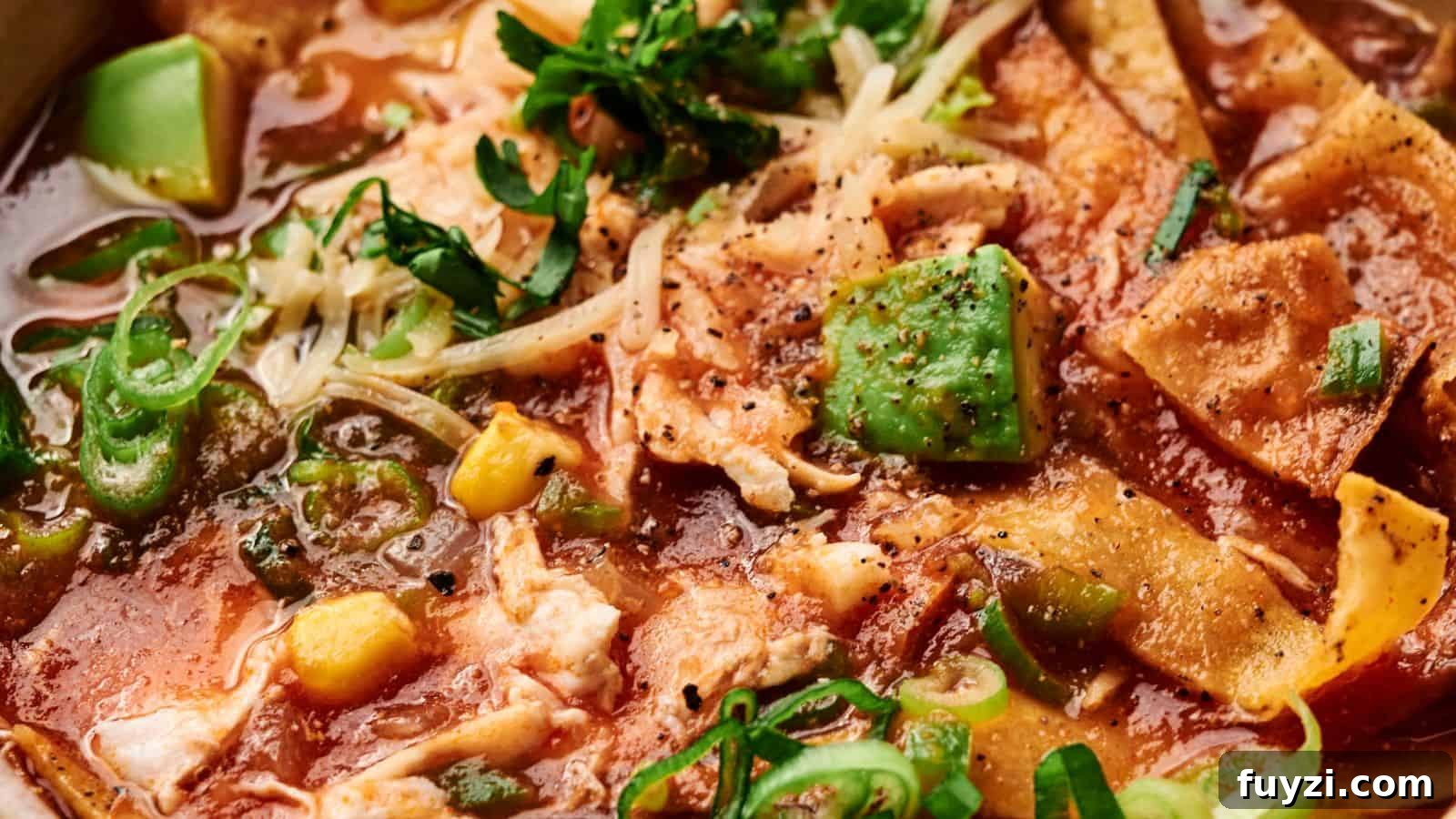 Close-up of a bowl of chicken tortilla soup with crispy tortilla strips, avocado pieces, corn, cheese, and herbs in a rich red broth.