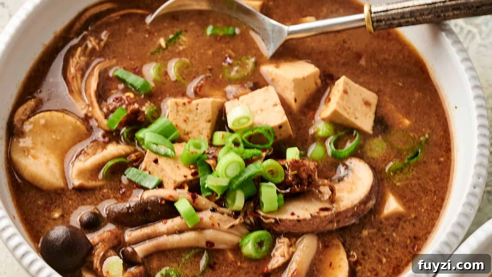 Bowl of hot and sour soup with mushrooms, tofu, and sliced green onions, with a spoon on the side.