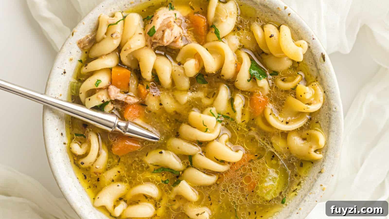 A bowl of rotini pasta soup with chunks of chicken, carrots, and herbs, with a spoon resting inside.