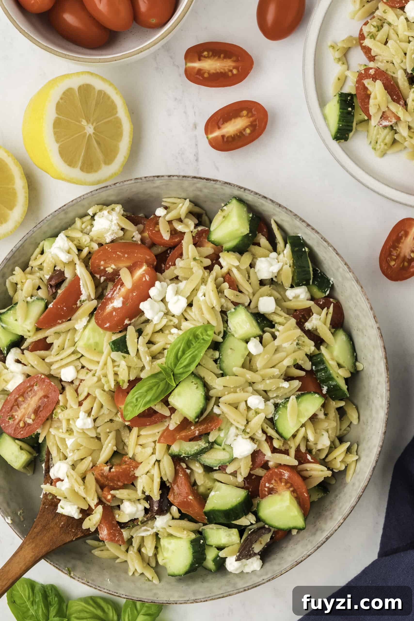 Creamy Pesto Orzo Pasta Salad with Cherry Tomatoes 2 A serving bowl with pasta salad with tomatoes and cucumbers. with a wooden spoon, some halved lemons and tomatoes found on the sides.