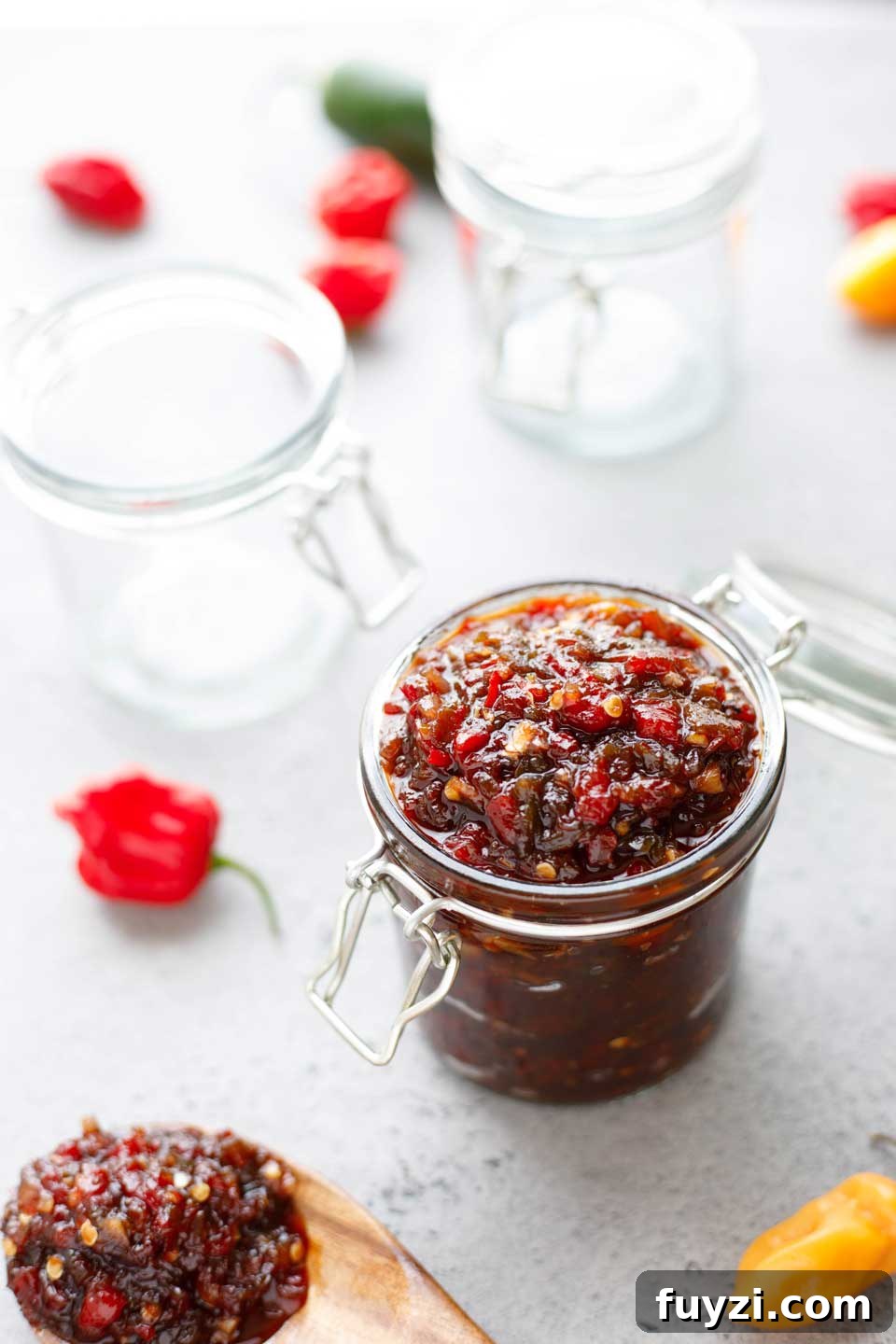 Glass jar filled with Pepper Relish, with a wooden spoonful, empty jars and a few hot peppers nearby.