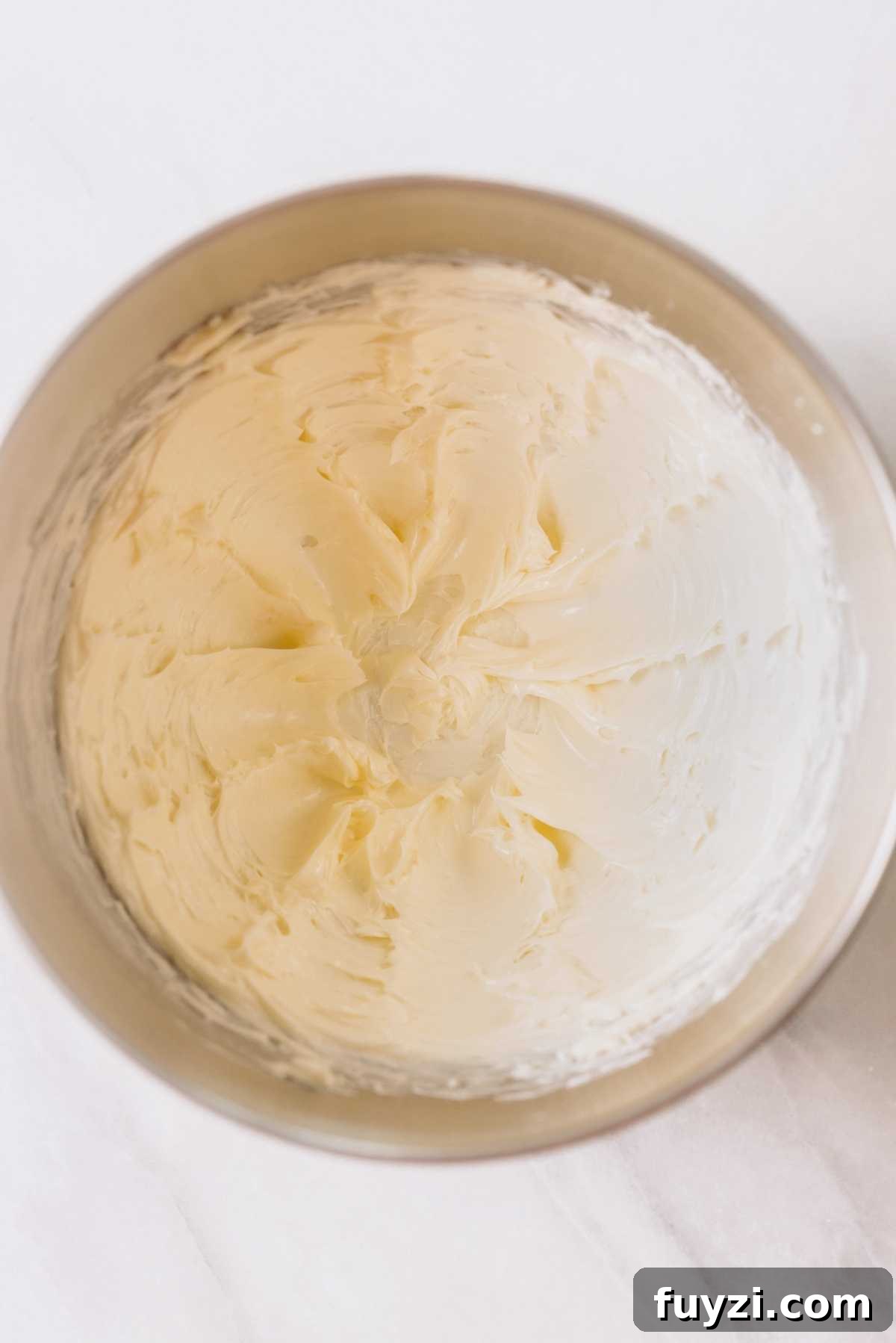 Unsalted butter creamed in a large metal mixing bowl on a white marbled background.