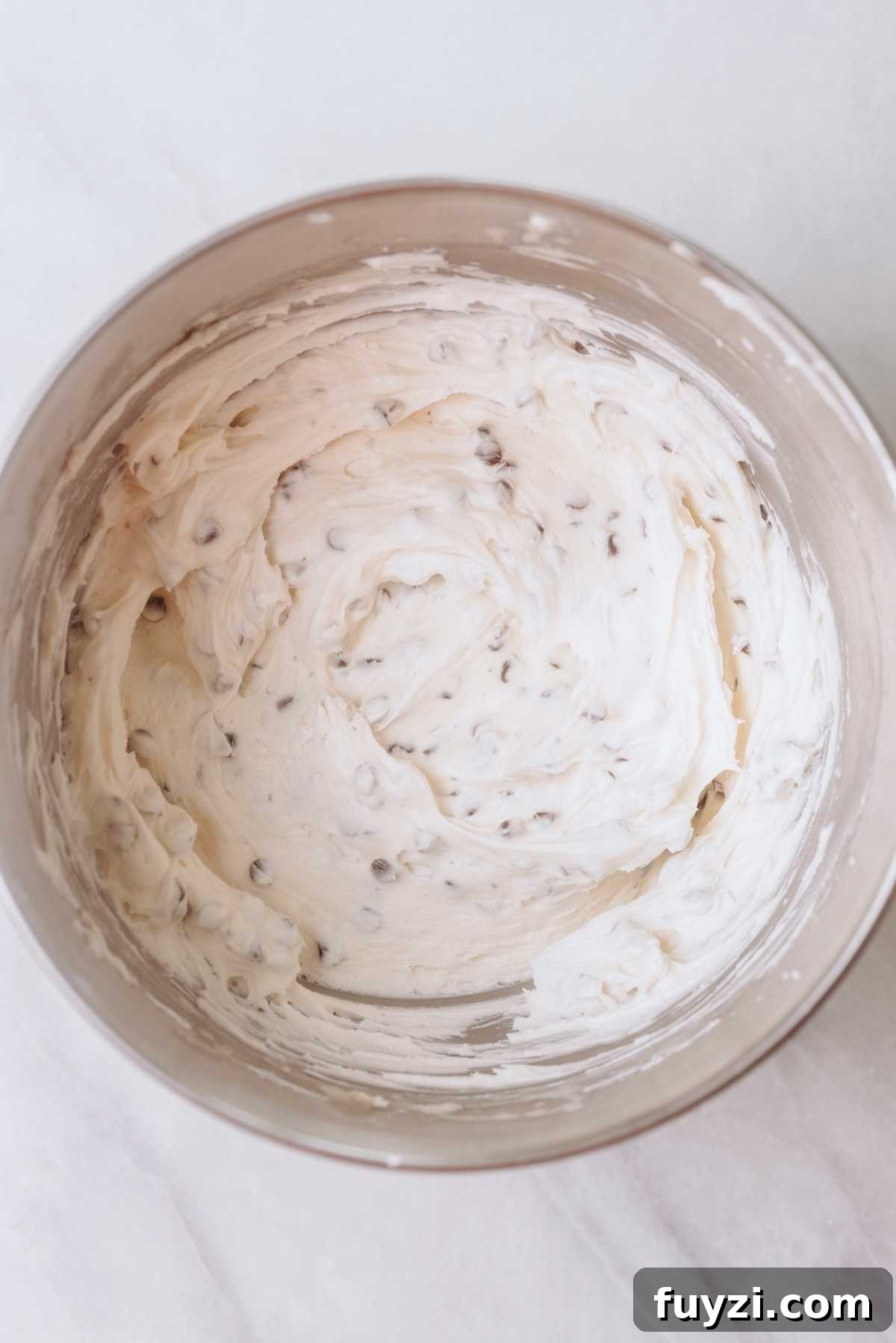 Chocolate chip buttercream frosting in a stainless steel mixing bowl on a white marbled background.
