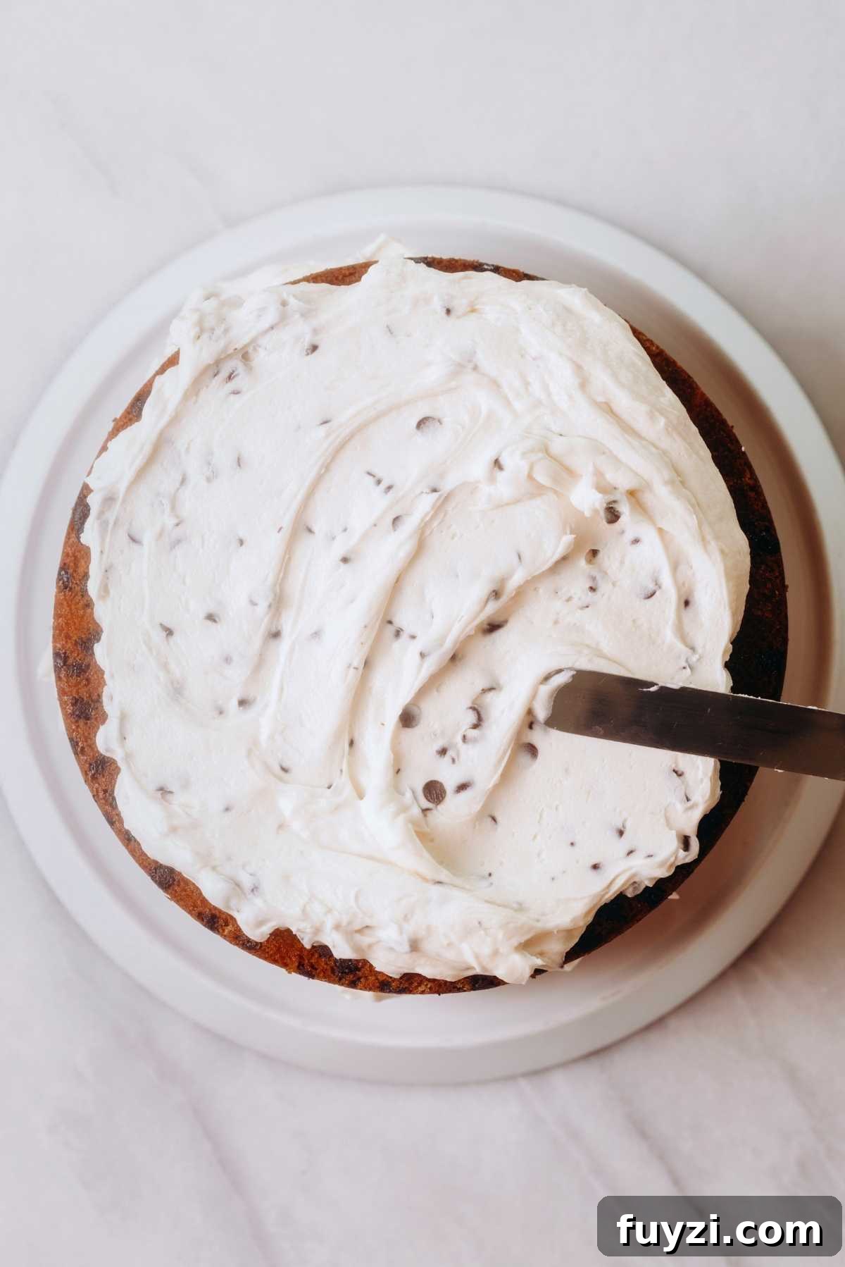 Frosting chocolate chip cake layers with chocolate chip buttercream frosting using an offset spatula.