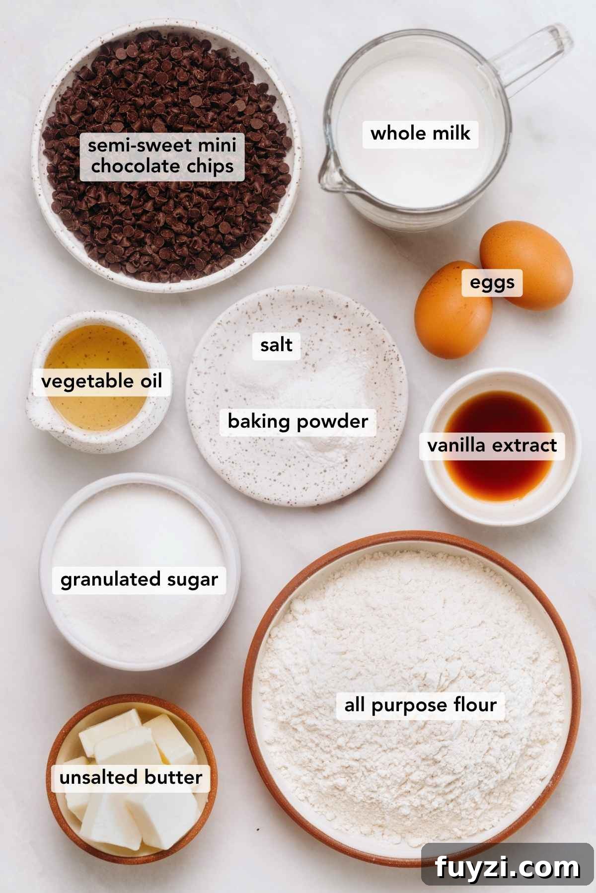 Ingredients in various size bowls labeled as semi sweet mini chocolate chips, whole milk, eggs, vegetable oil, salt, baking powder, vanilla extract, granulated sugar, all purpose flour, and unsalted butter on a white marbled background.