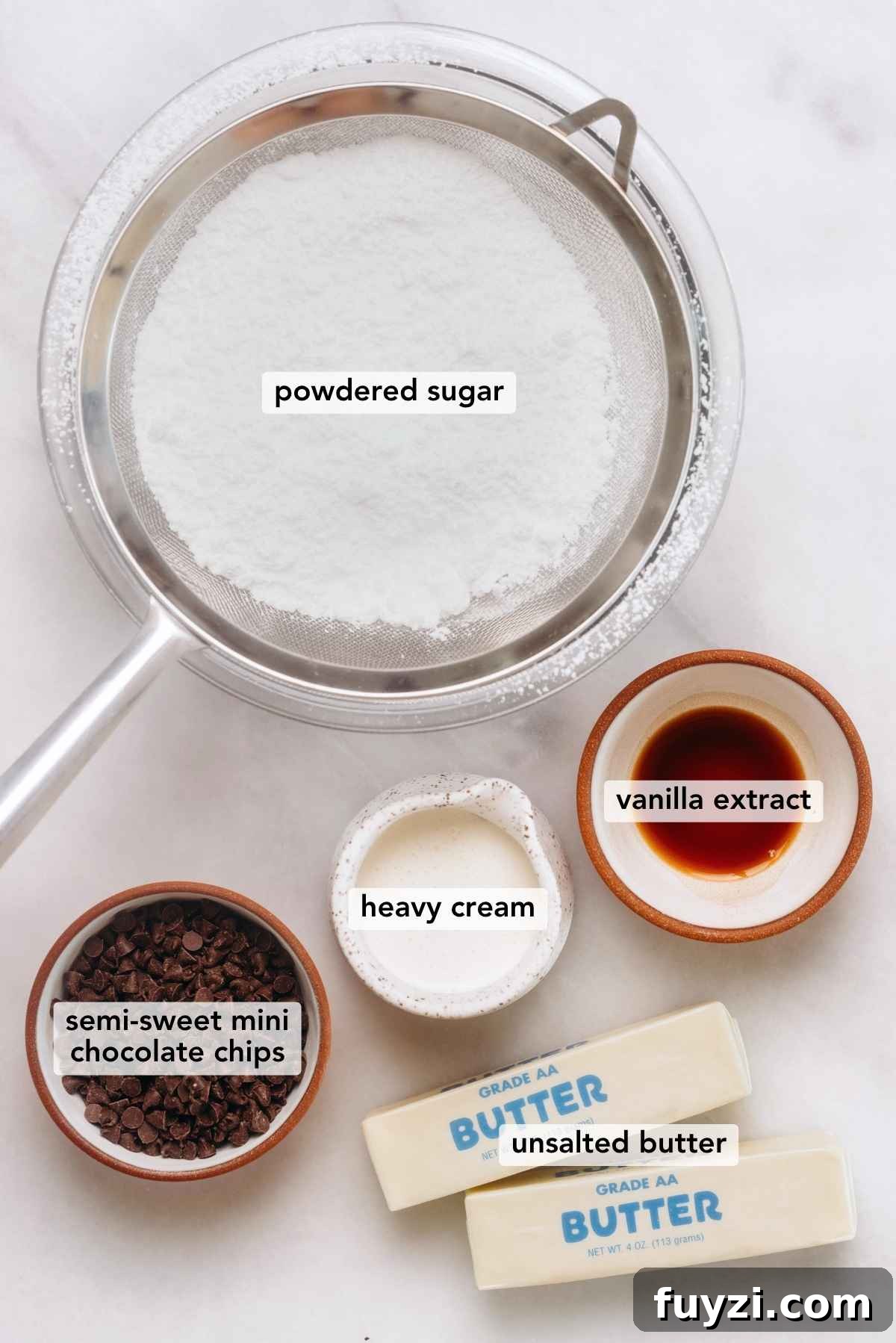 Ingredients in various size bowls labeled as powdered sugar, semi sweet mini chocolate chips, vanilla extract, heavy cream, and unsalted butter on a white marbled background.