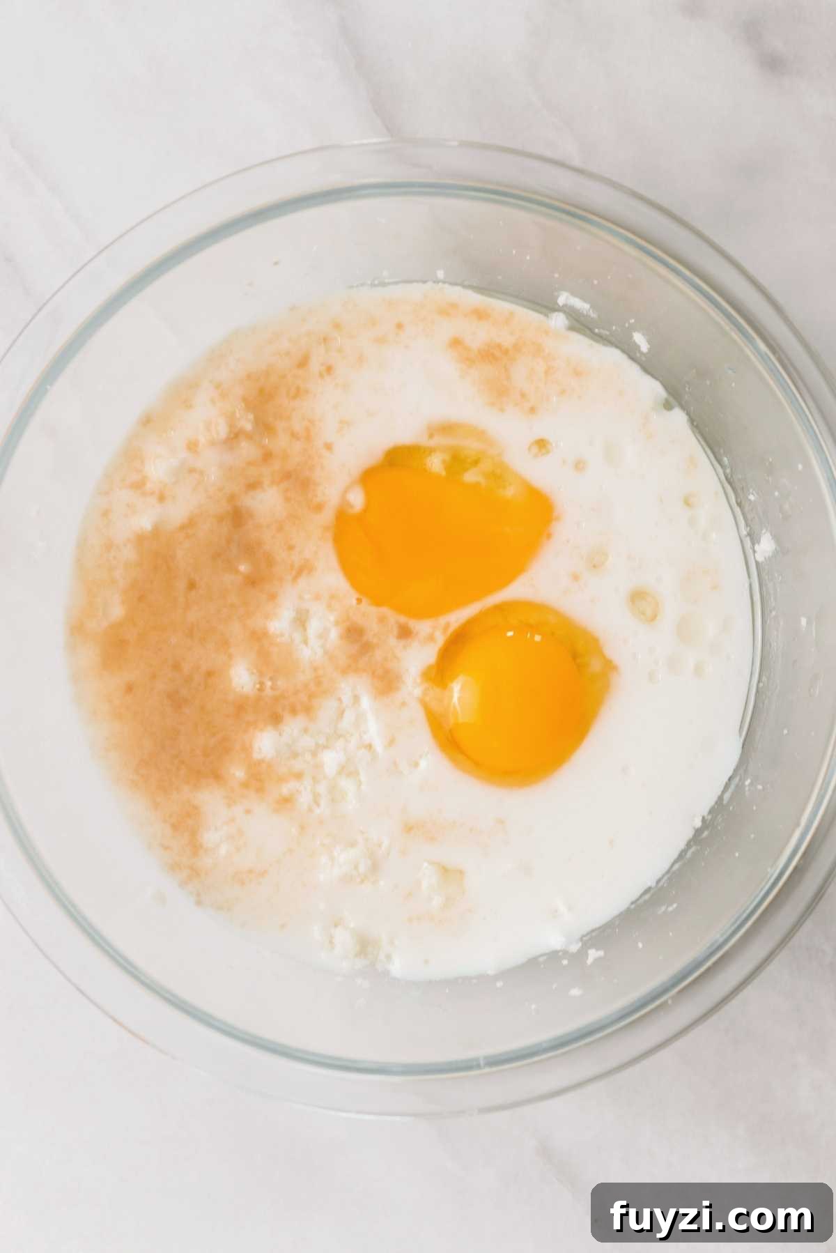 Unsalted butter and sugar creamed together in a large glass mixing bowl with eggs, oil, vanilla extract, and milk added on a white marbled background.