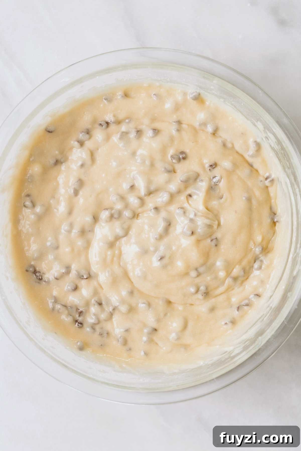 Chocolate chip cake batter in a large glass mixing bowl on a white marbled background.