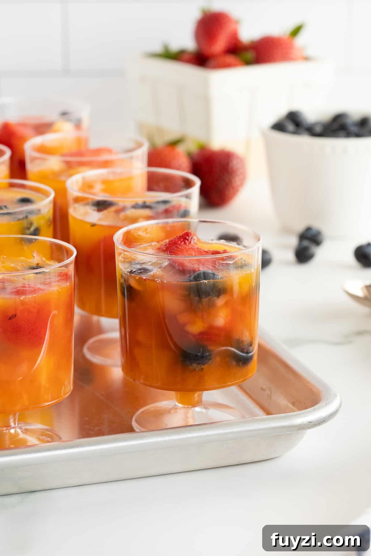 A close up of plastic cups filled with frozen fruit on a baking sheet next to bowls of strawberries and blueberries.