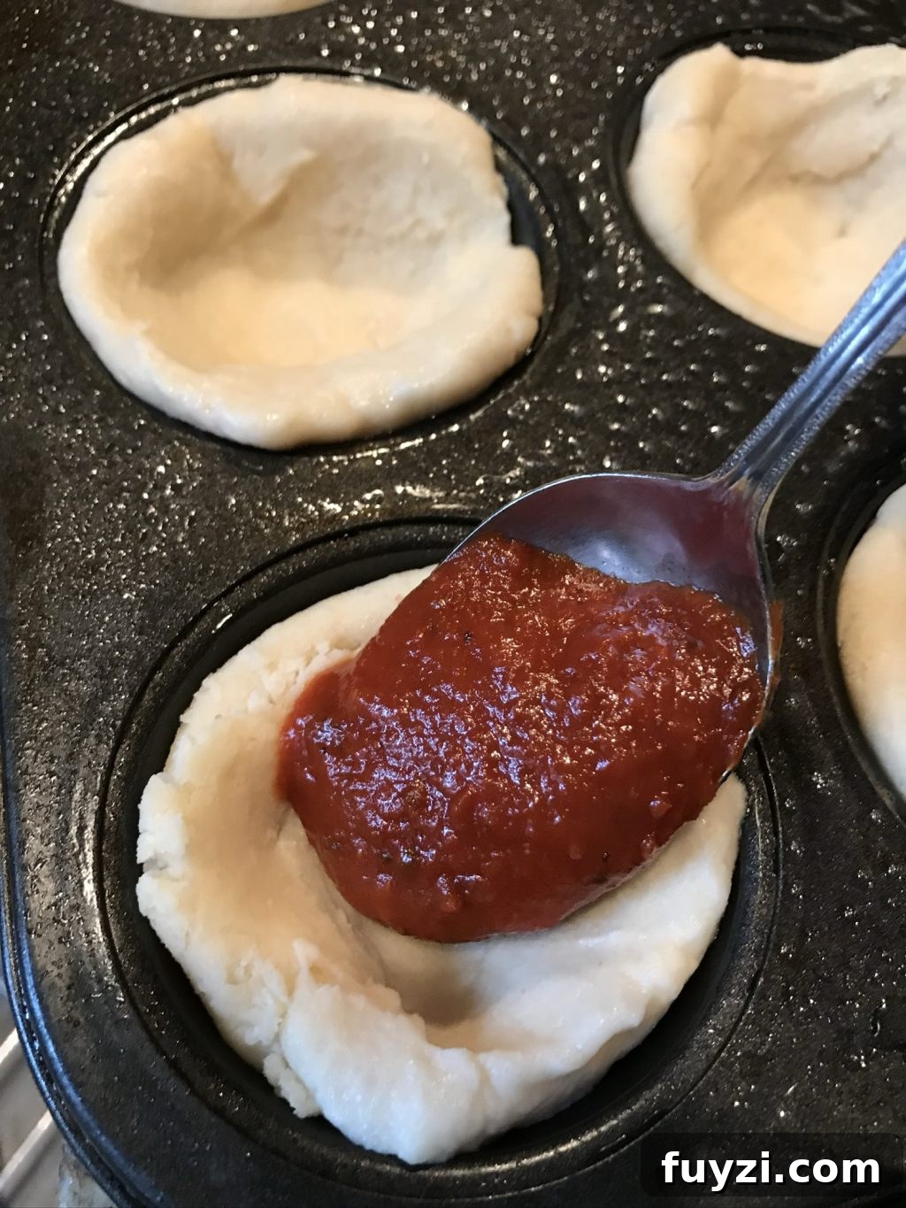 Pizza sauce being spooned into one of the dough filled muffin tin cups.