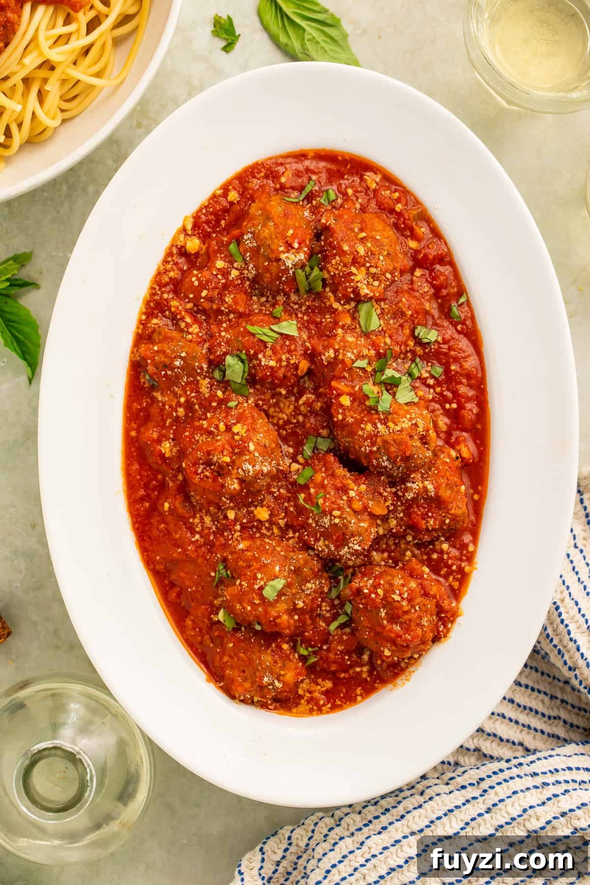 White oval serving platter of meatballs in tomato sauce. A bowl of cooked spaghetti and bottle of olive oil is visible off to one side.