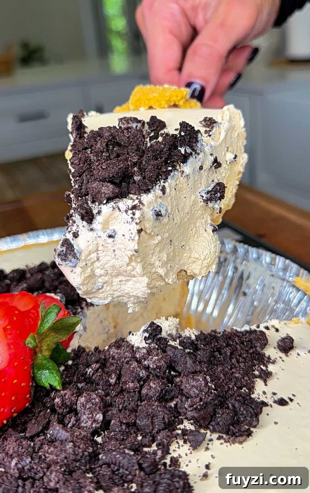 No-Bake Vintage Coffee Ice Cream Pie Recipe 2 A hand lifts a large, creamy slice of pie topped with crushed chocolate cookies from a pie tin. A fresh strawberry sits beside the pie, and the background shows a kitchen setting.
