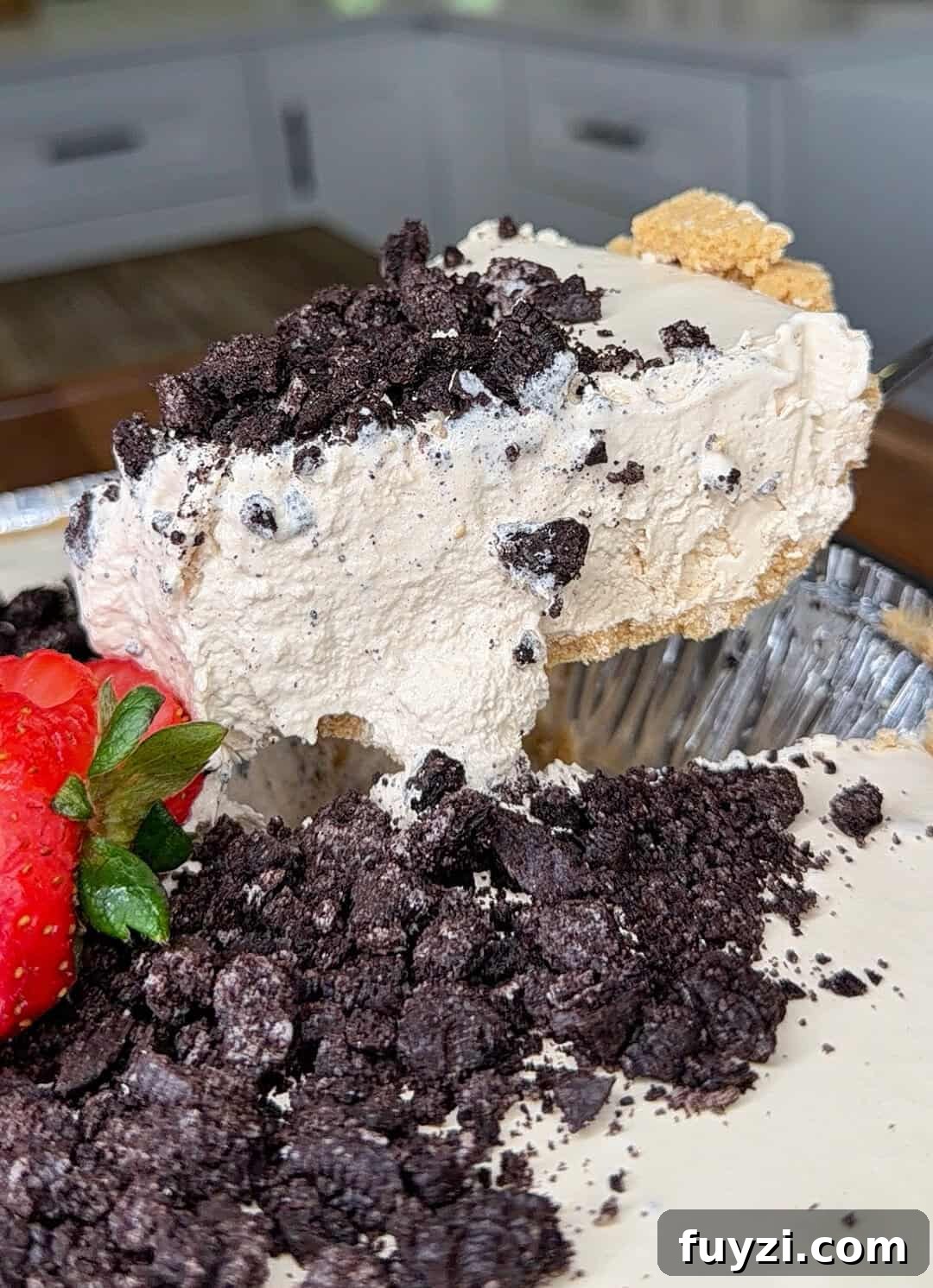 No-Bake Vintage Coffee Ice Cream Pie Recipe 8 A close-up of a creamy pie slice topped with crushed chocolate cookies and a fresh strawberry, set in a metal pie tin on a kitchen counter.