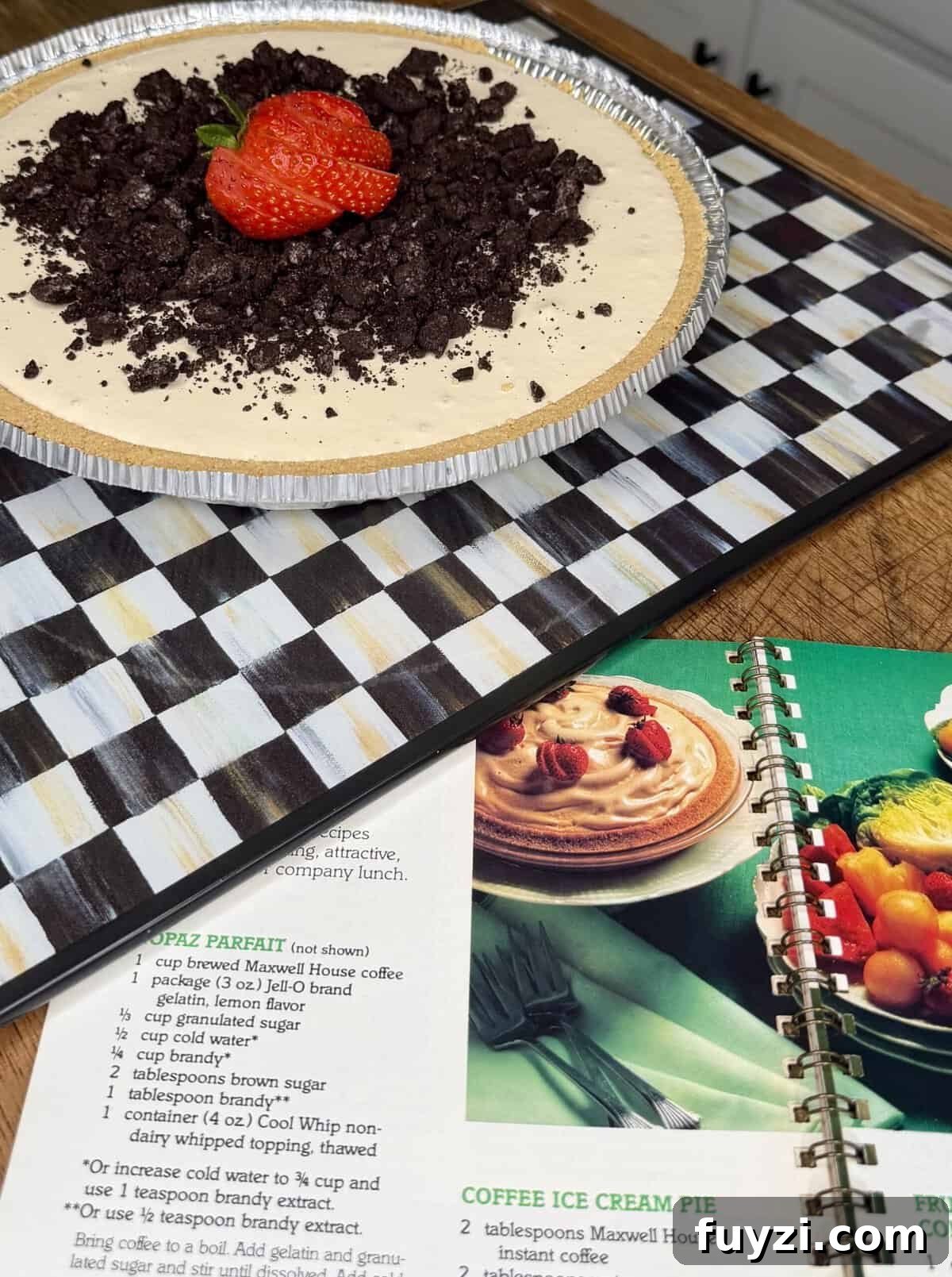 No-Bake Vintage Coffee Ice Cream Pie Recipe 7 A creamy pie with crumbled chocolate cookies and a strawberry on top sits on a checkered board next to an open cookbook displaying a similar dessert recipe.