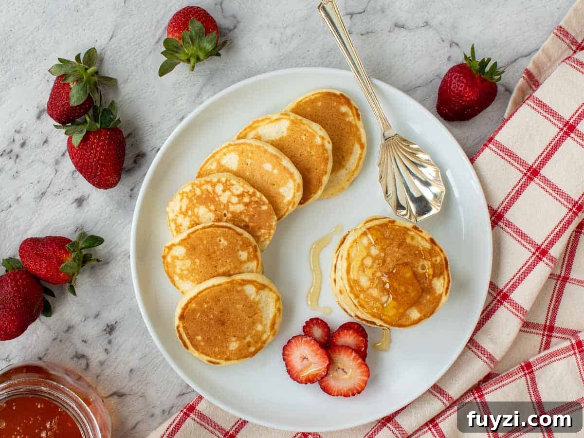 Four pancakes stacked on a white plate with syrup dripping off and more pancakes and strawberries around viewed from above.