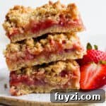 A stack of three strawberry rhubarb bars on a plate with two fresh strawberries next to them.