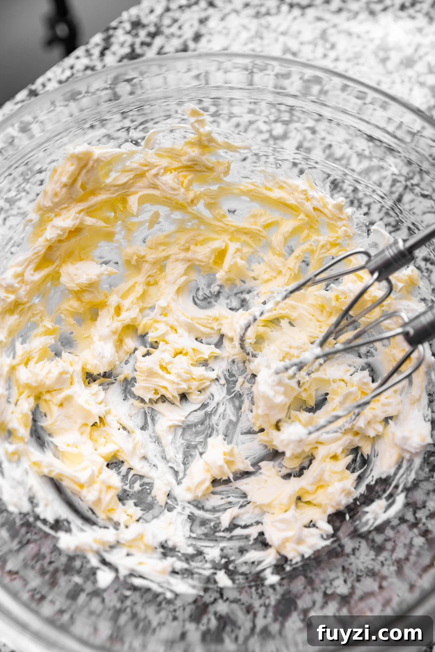 Cream cheese and butter being beaten together in a glass mixing bowl.