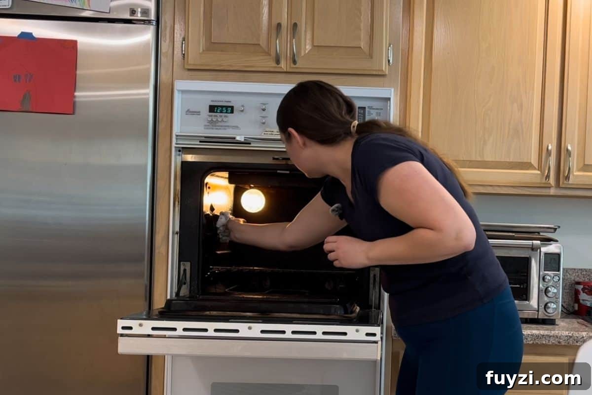 A woman wearing a navy shirt and blue pants is wiping out her oven in a kitchen with wooden cabinets.
