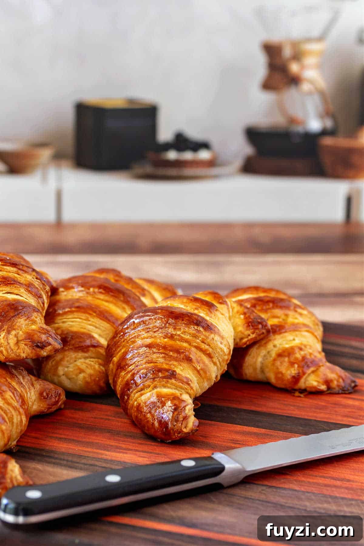 Several shiny and deep golden brown croissants on a cutting board with a bread knife.