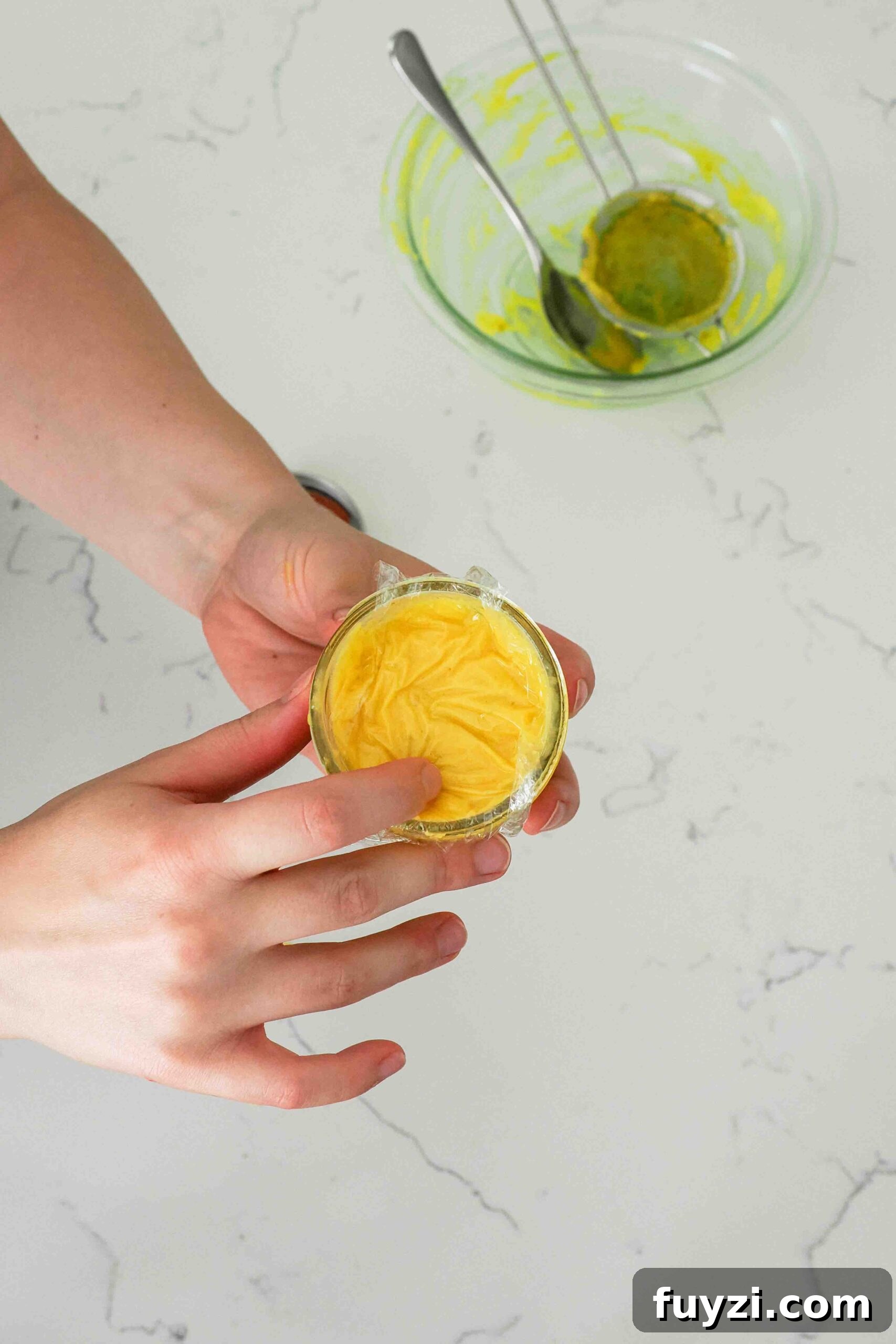 A hand presses plastic wrap to the top of a jar of mango curd.