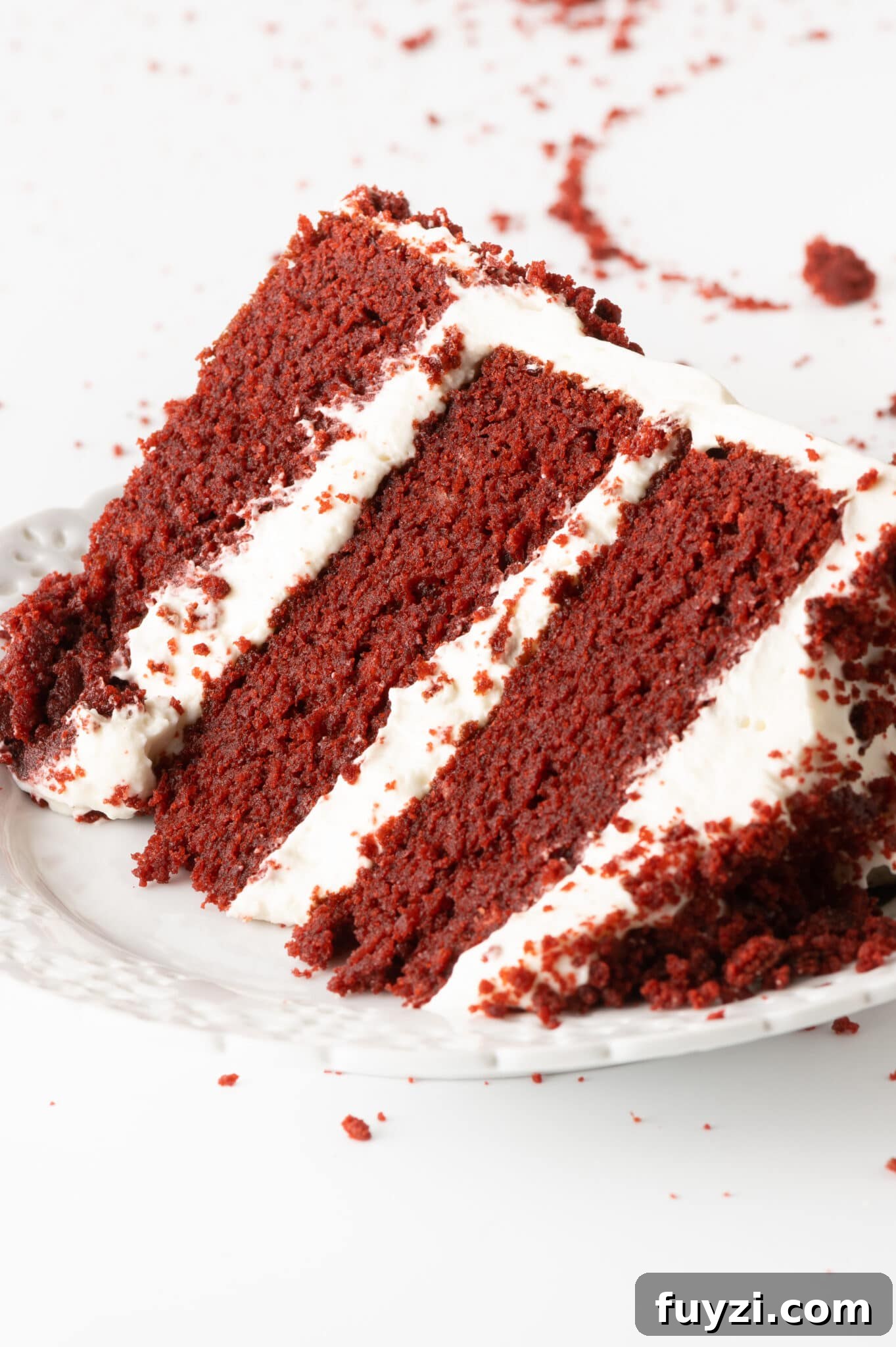 Low-Carb Red Velvet Cake Recipe with Cream Cheese Frosting 16 A single slice of bright red maroon red velvet cake with bright white layers of frosting. The cake is set on a white texture plate and set against a bright white tabletop with red cake crumbs scattered around the plate.