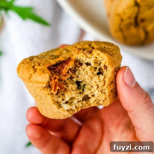 A hand holding a savoury chickpea flour muffin - a bite has been taken revealing the fluffy texture and a sun-dried tomato. There are more muffins on a plate in the background.