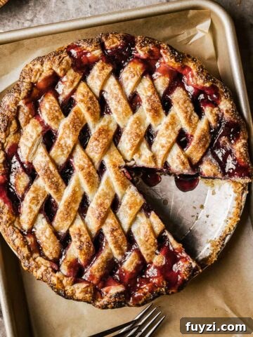 A golden brown lattice topped baked cherry rhubarb pie on a parchment lined baking sheet.