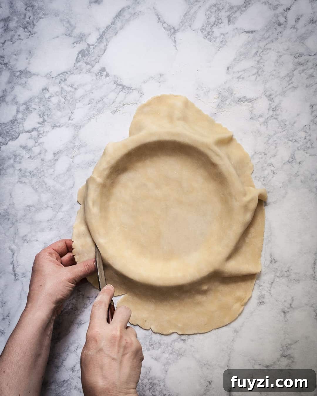 Hands trimming excess pie dough around a pie pan.