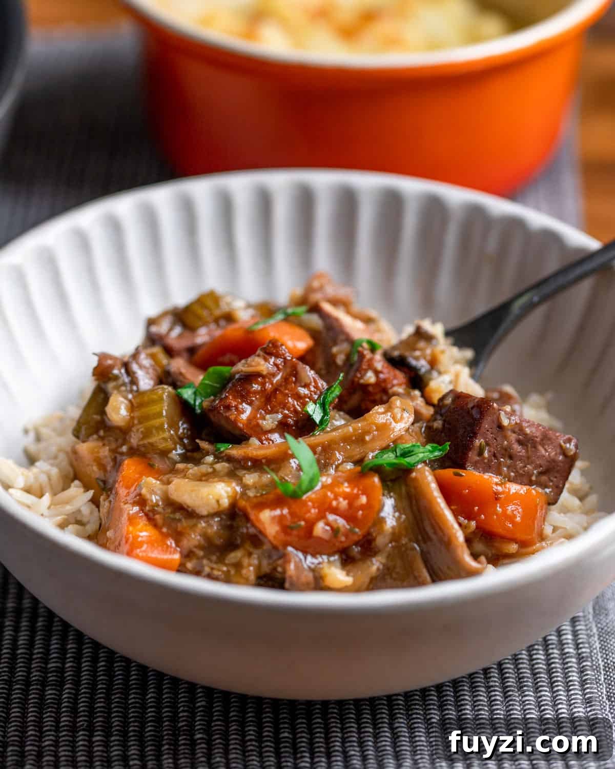 Vegan Smoked Tofu and Vegetable Stew 3 Photo of a bowl of vegan stew served with brown rice and a spoon, set on a grey mat with an orange pot in the background.