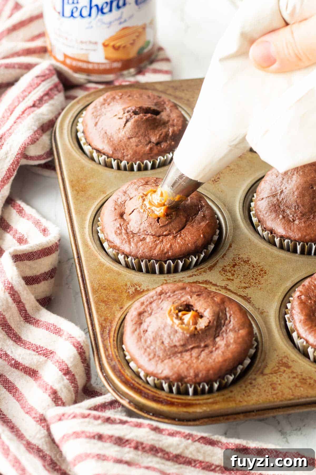 Gooey Chocolate Caramel-Filled Cupcakes Recipe 6 Hand piping dulce de leche into baked chocolate cupcakes in metal pan.