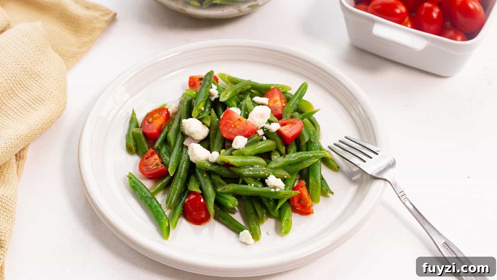 27 Crowd-Pleasing Outdoor Potluck Recipes for Late Summer 17 A plate of green beans tossed with cherry tomatoes and crumbled feta cheese, accompanied by a fork, on a white surface.
