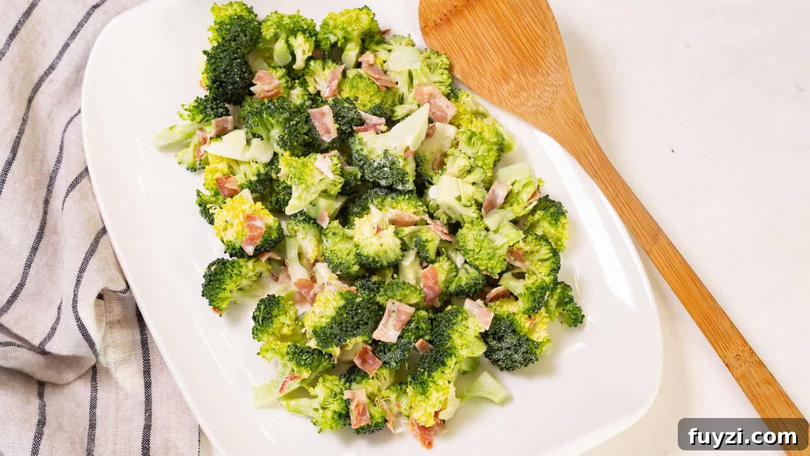 27 Crowd-Pleasing Outdoor Potluck Recipes for Late Summer 18 Broccoli salad with bacon bits on a white oval plate, accompanied by a wooden serving spoon on a linen napkin.