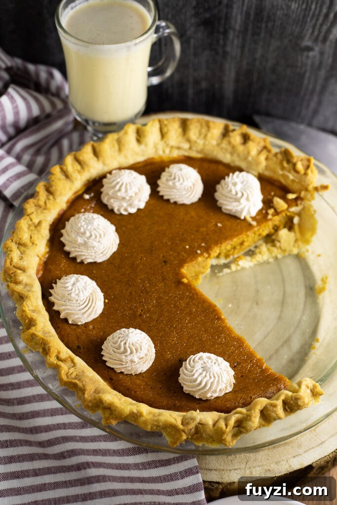 An eggnog pumpkin pie in a glass pie dish topped with piped swirls of whipped cream and a large slice missing. A glass of eggnog and a linen napkin are in the background.