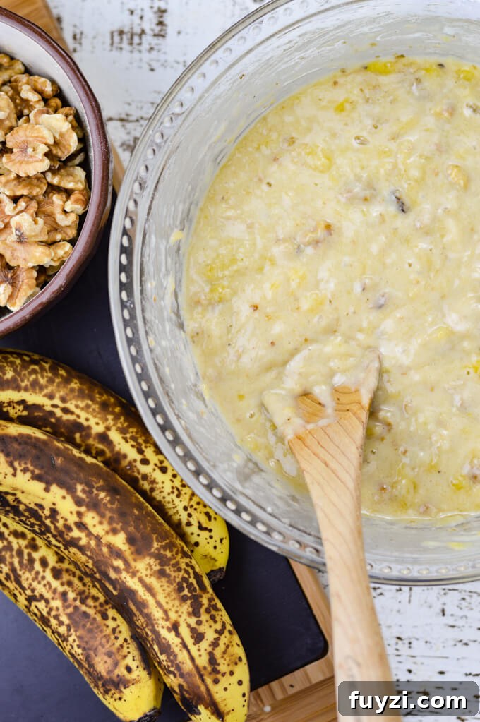 Moist Banana Nut Bread Recipe for Tender, Flavorful Loaves 5 A bowl of banana nut bread batter with a wooden spoon in it. Ripe bananas and a bowl of walnuts sit next to it.