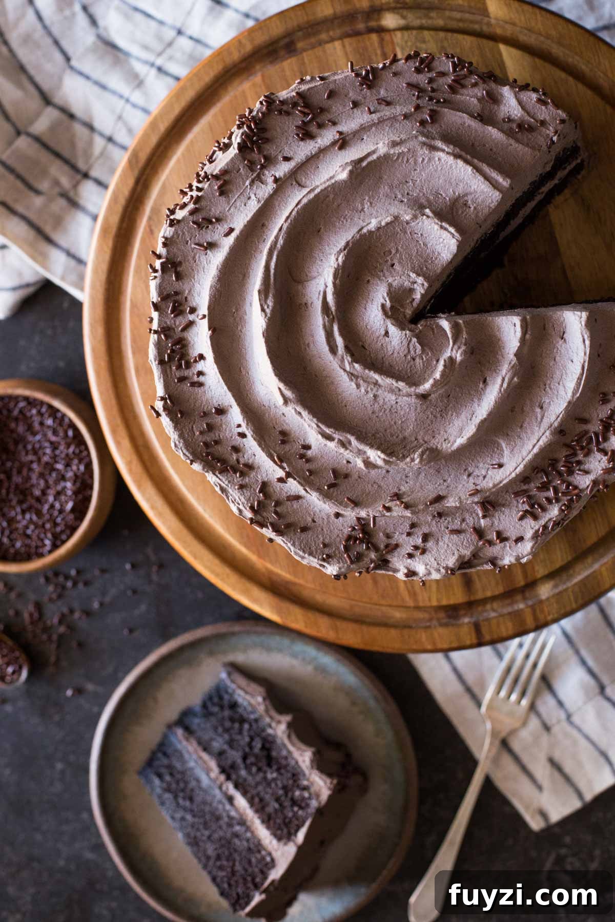 A slice of Dark Chocolate Cake With Whipped Cream Frosting on a plate sitting next to the rest of the cake on a cake stand and a small wood bowl of chocolate sprinkles. 