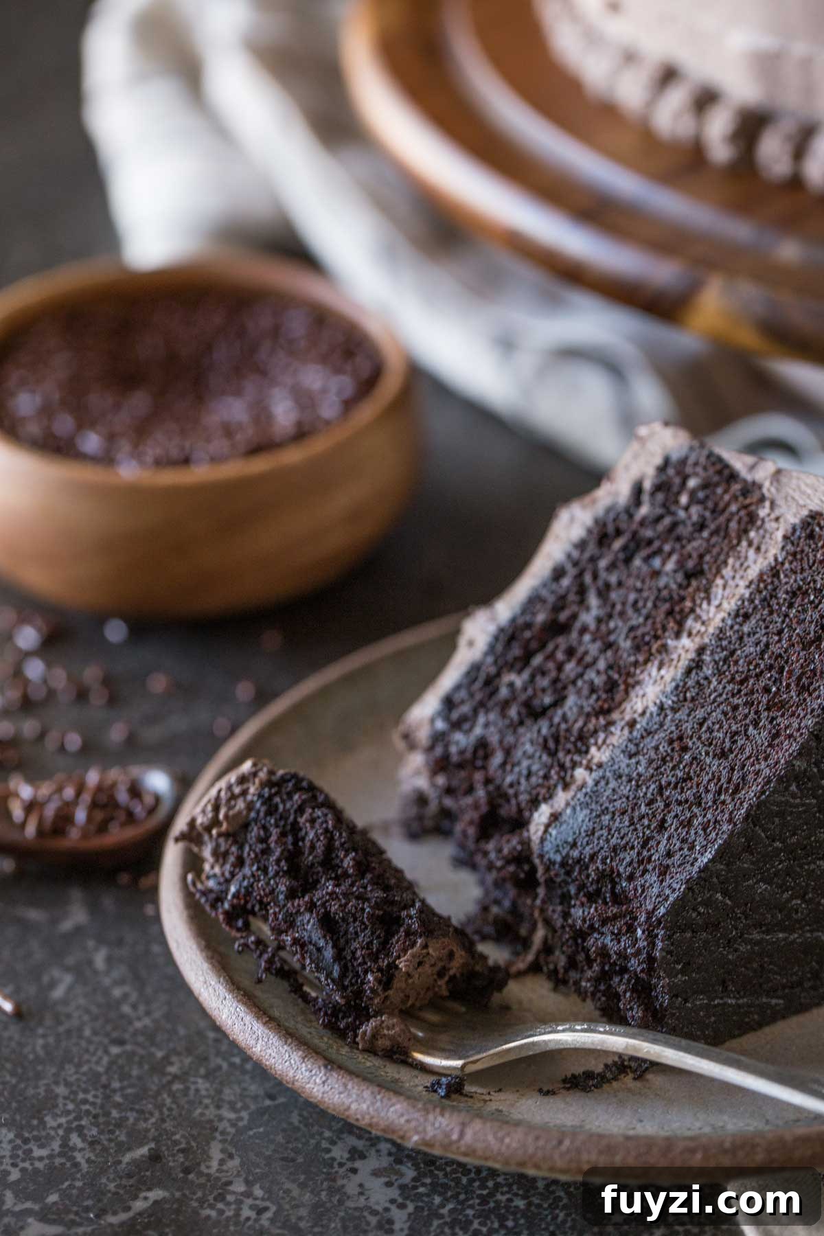 A slice of Dark Chocolate Cake With Whipped Cream Frosting on a plate along with a fork full of cake, and a small wood bowl of chocolate sprinkles in the background. 